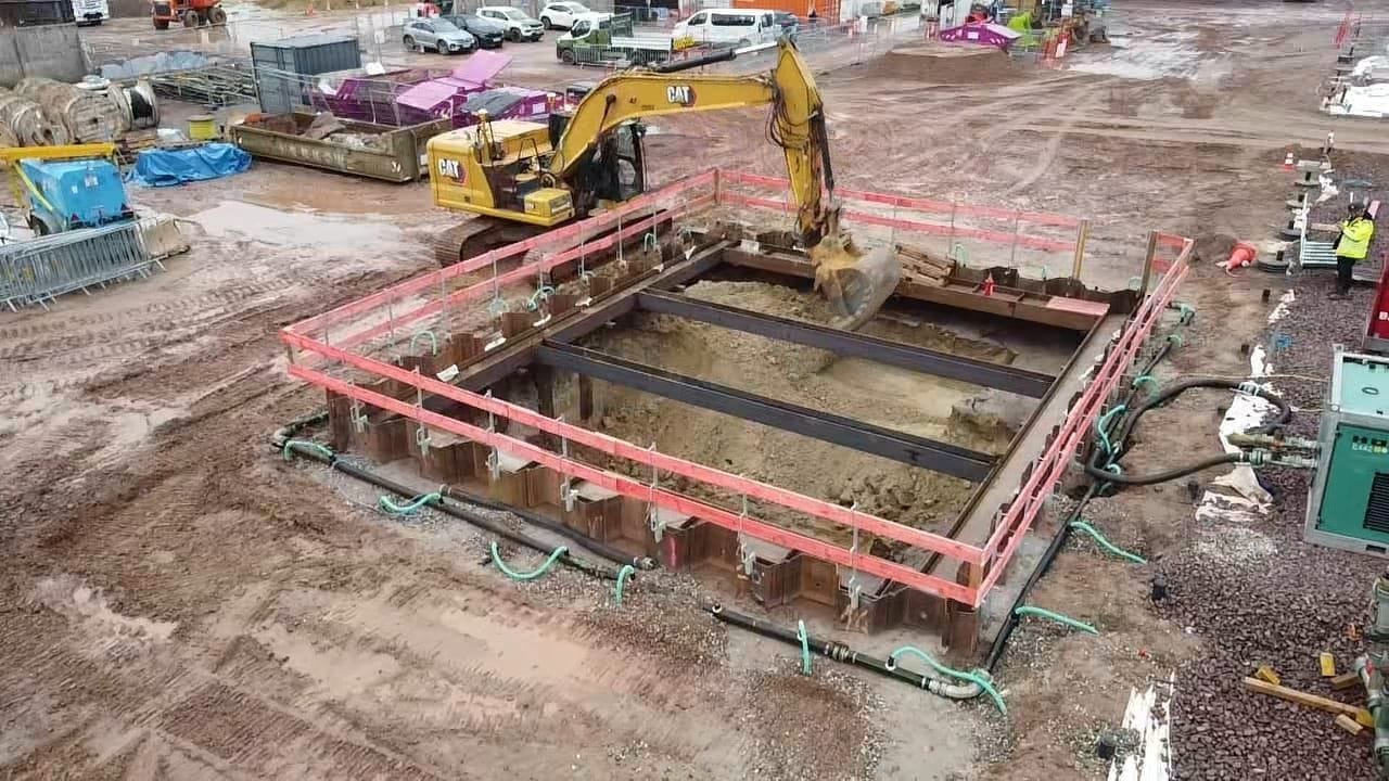 A Maveric excavator digging a shored, dewatered foundation within sheet-piled support on a live construction site.