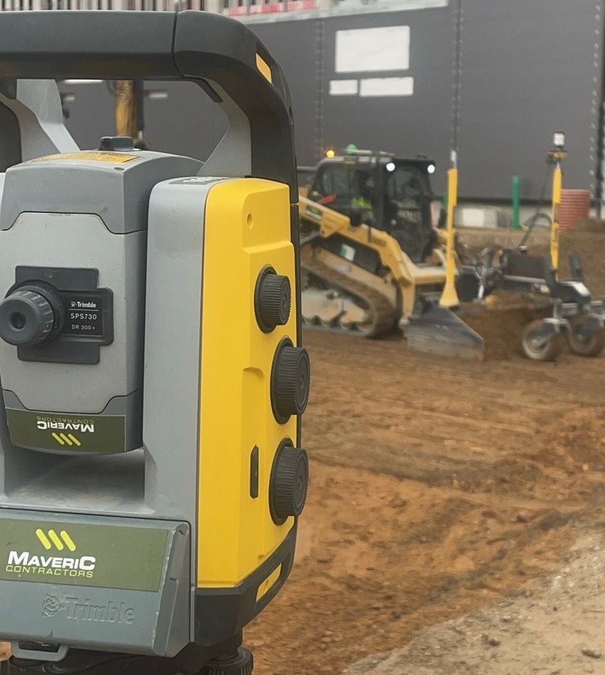A Maveric-branded Trimble SPS730 total station on a tripod, with a tracked loader grading the platform behind it.